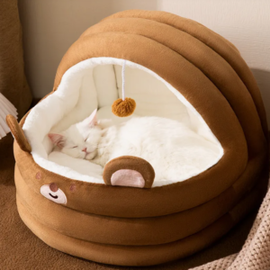 White cat sleeping in cozy brown plush cat bed with bear face design and hanging toy.