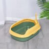 Open green and yellow cat litter box with scoop on gray tile floor near plant